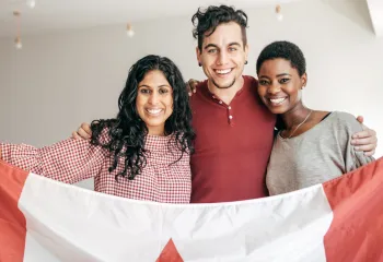 A man and two women, all of different cultural backgrounds, smiling and holding up a Canadian flag together
