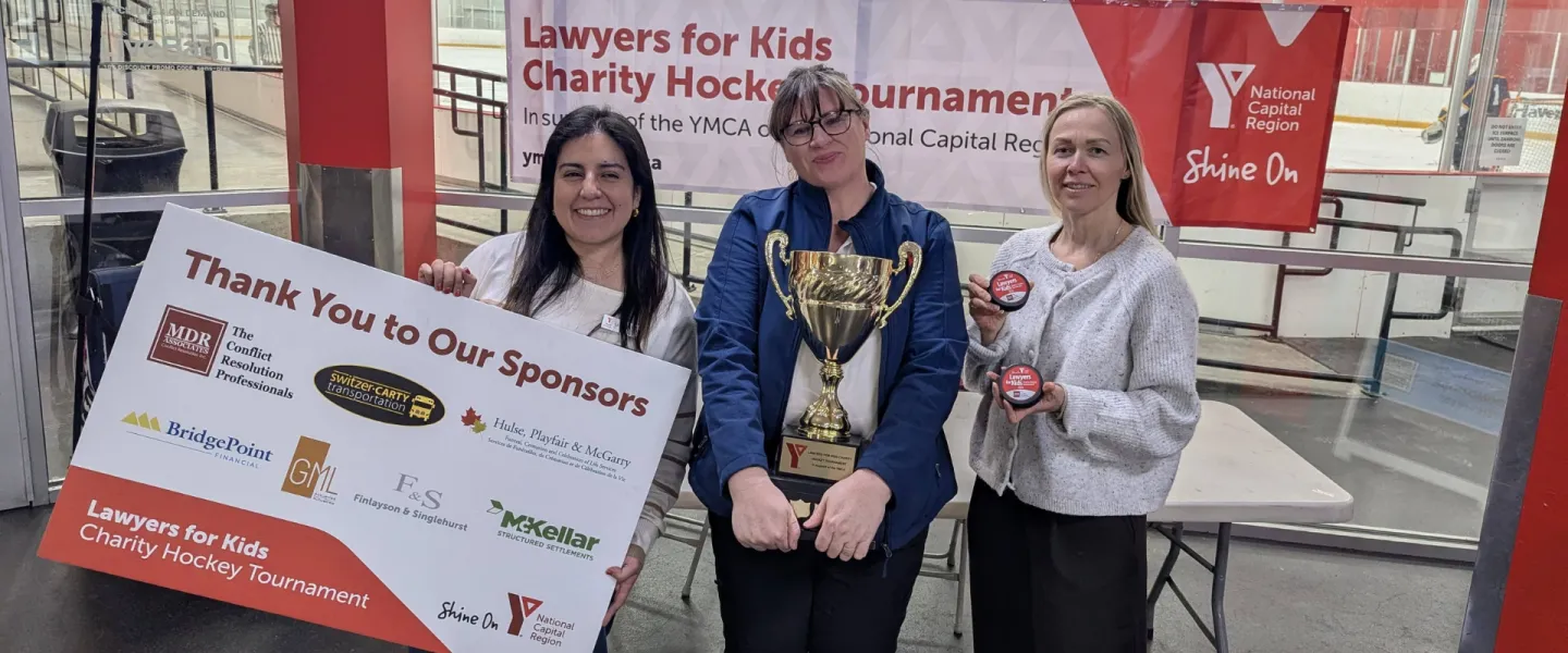Maria, Marianne, and Natalya from the YMCA Philanthropy team holding up a "thank you to our sponsors" logo board, a golden trophy, and hockey pucks.