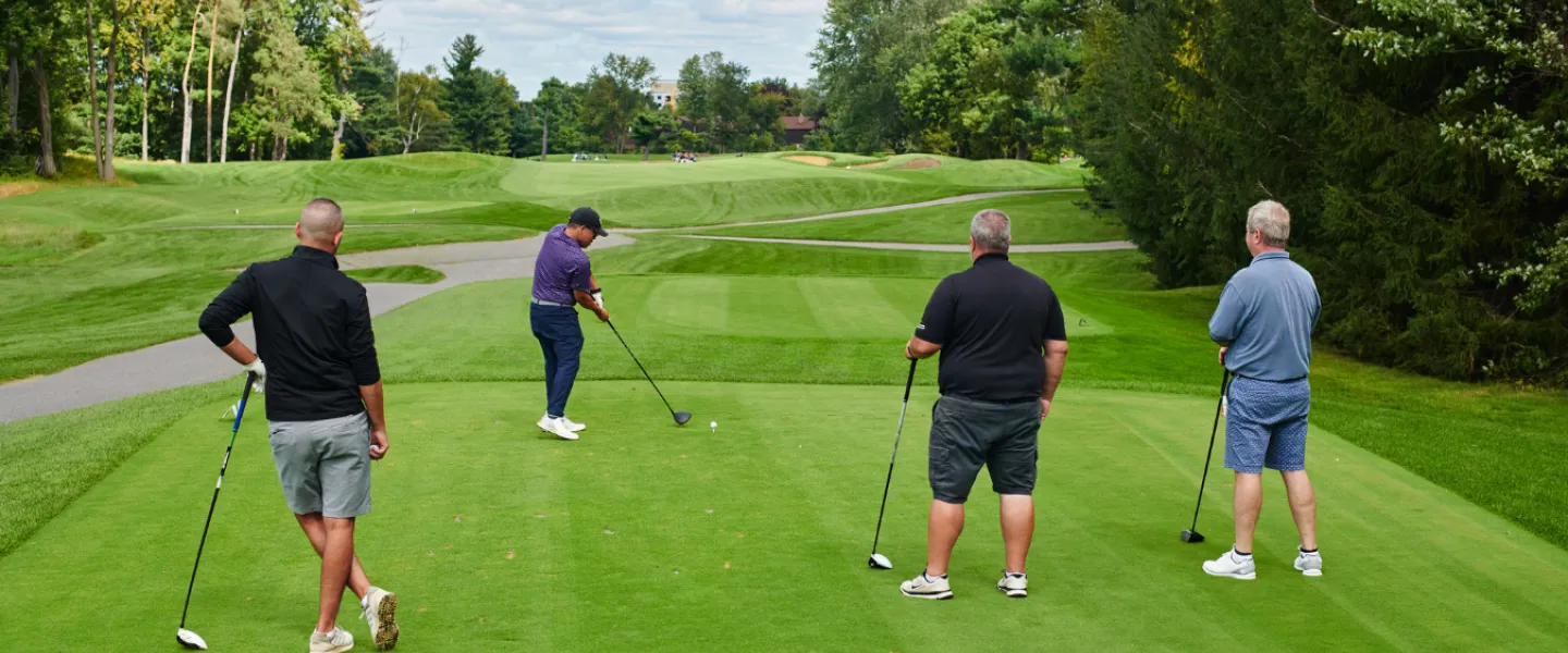 A team of four golfers on a course. One golfer is teeing up.